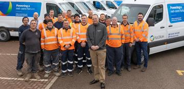 Image shows 20 men in workwear standing in front of several leased vans, in a car park. The majority in high visibility, orange shirts. Vans have Fulcrum branding and blue graphic stating 'powering a net-zero future'.