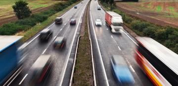 Duel carriageway between two fields. Images shows cars and haulage vehicles that are blurred, moving at speed, in both directions.