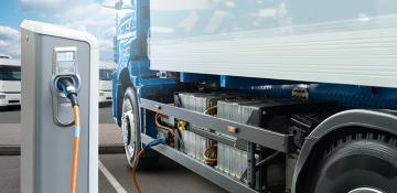 Image shows a long wheelbase van plugged into an electric charging point. The charger in in the foreground, with an orange cable plugged in leading to the white van which has a blue tractor